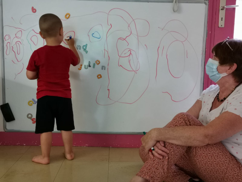 Un enfant dessine sur un tableau blanc avec des feutres colorés, une femme le regarde.